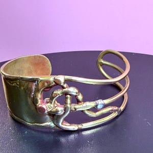 May include: A gold-toned cuff bracelet with an open design. The bracelet features a wide band and intricate, intertwined metalwork with a small, light-colored stone accent. The overall style is artistic and unique.