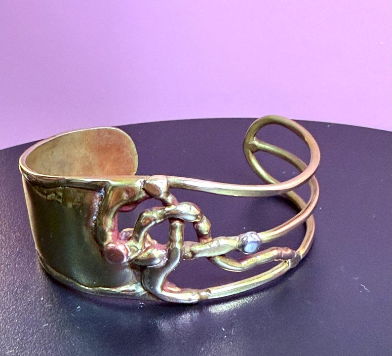 May include: A gold-toned cuff bracelet with an open design. The bracelet features a wide band and intricate, intertwined metalwork with a small, light-colored stone accent. The overall style is artistic and unique.