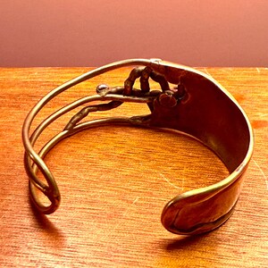 May include: A handcrafted copper cuff bracelet with an open design. The bracelet features a detailed, stylized hand grasping a small, clear bead. The cuff has a curved shape and a polished finish, showcasing the warm tones of the metal.