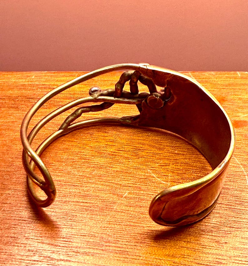 May include: A handcrafted copper cuff bracelet with an open design. The bracelet features a detailed, stylized hand grasping a small, clear bead. The cuff has a curved shape and a polished finish, showcasing the warm tones of the metal.