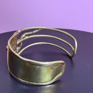 May include: A gold-toned cuff bracelet with a wide, curved band and three parallel, thinner bands. The bracelet has a textured design on the side and a small, dark accent. The jewelry is displayed against a purple background.