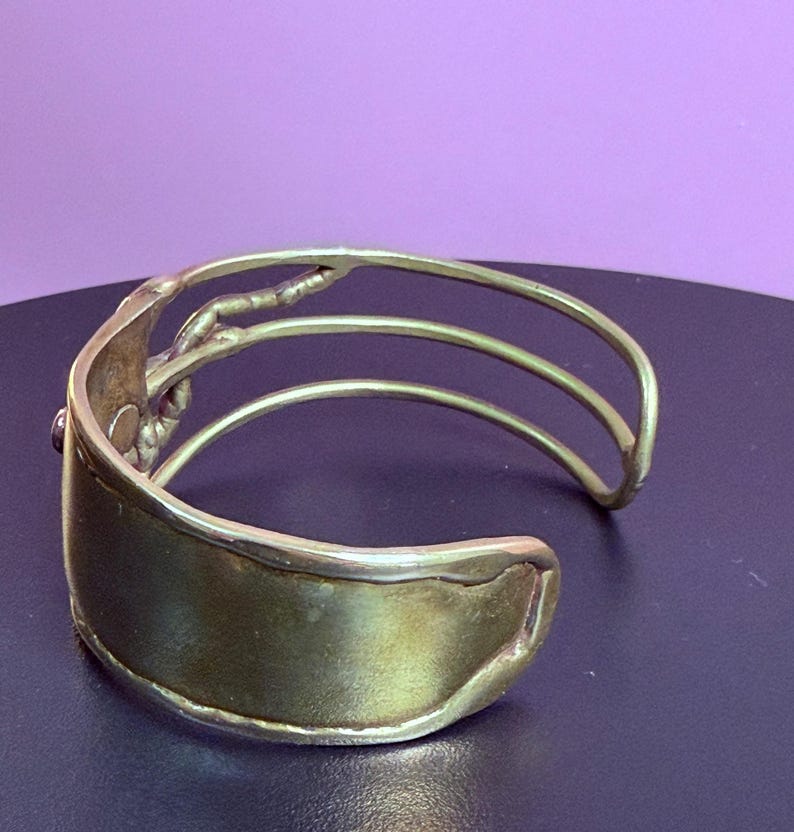 May include: A gold-toned cuff bracelet with a wide, curved band and three parallel, thinner bands. The bracelet has a textured design on the side and a small, dark accent. The jewelry is displayed against a purple background.