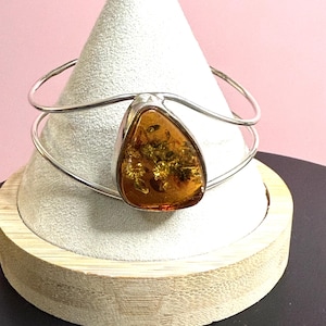 May include: A silver cuff bracelet with a large, amber stone. The amber is a warm, golden-brown colour with visible inclusions. The bracelet is displayed on a white cone-shaped stand with a wooden base, against a pink backdrop.