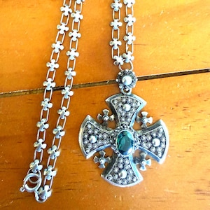 May include: A silver-toned metal necklace featuring a cross pendant with intricate filigree detailing and a central oval-shaped dark green gemstone. The chain is composed of repeating small cross-shaped links, and the pendant includes a small circular accent at the top.