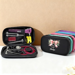 Könnte beinhalten: Ein schwarzes medizinisches Etui mit einem roten Stethoskop und medizinischen Werkzeugen. Das Etui ist geöffnet und zeigt das Stethoskop und andere medizinische Instrumente. Ein schwarzes Etui mit einer rosa Schleife und dem Namen "Penelope" ist ebenfalls zu sehen.