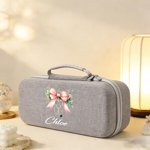 Könnte beinhalten: Graues medizinisches Etui mit rosa Schleife und Stethoskop-Design, mit dem Namen "Chloe" in weißer Schrift. Das Etui hat einen Griff und einen Reißverschluss. Das Design beinhaltet florale Akzente.