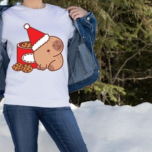 May include: White sweatshirt featuring a cartoon sloth wearing a Santa hat, sitting next to a red cookie tin. Several chocolate chip cookies are scattered around the tin. The design is in a cartoon style, perfect for the holiday season.