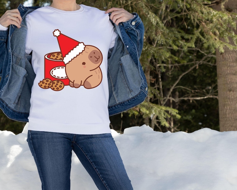 May include: White sweatshirt featuring a cartoon sloth wearing a Santa hat, sitting next to a red cookie tin. Several chocolate chip cookies are scattered around the tin. The design is in a cartoon style, perfect for the holiday season.