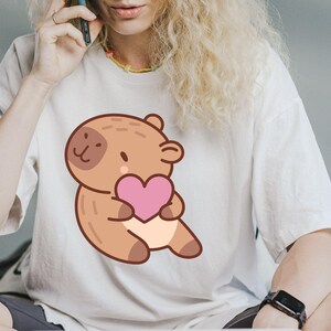 May include: White t-shirt featuring a cartoon capybara holding a pink heart. The capybara is brown with a light beige belly and a dark brown spot on its face. The shirt is a casual, oversized fit.