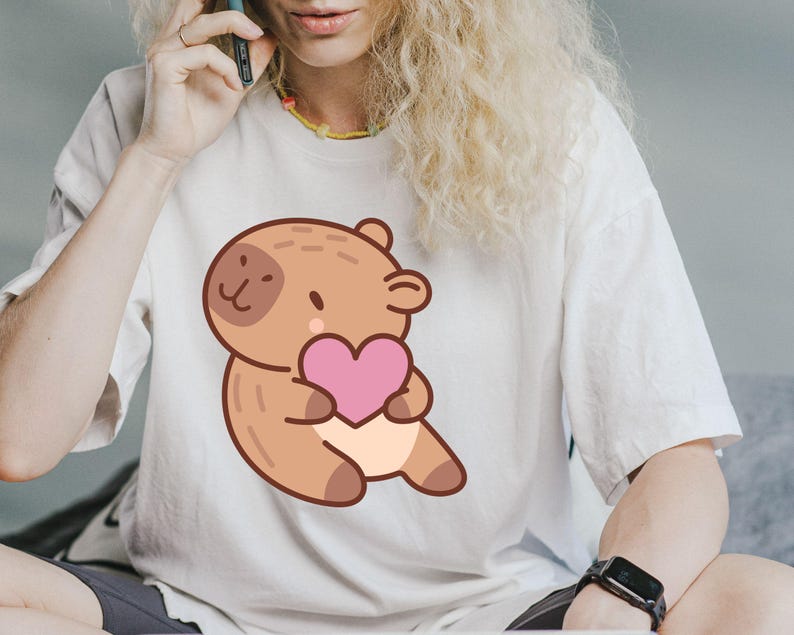May include: White t-shirt featuring a cartoon capybara holding a pink heart. The capybara is brown with a light beige belly and a dark brown spot on its face. The shirt is a casual, oversized fit.