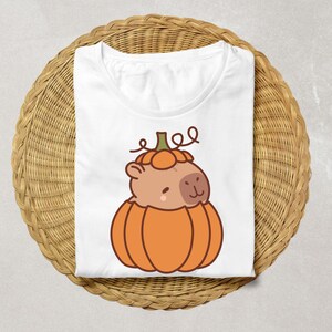 May include: White t-shirt featuring a cartoon capybara inside an orange pumpkin. The capybara has a light brown face and is smiling. The pumpkin has a green stem and is sitting on a woven wicker basket.