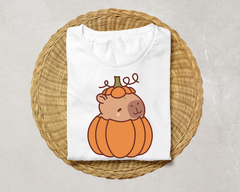 May include: White t-shirt featuring a cartoon capybara inside an orange pumpkin. The capybara has a light brown face and is smiling. The pumpkin has a green stem and is sitting on a woven wicker basket.