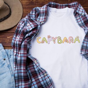 May include: White t-shirt with the word "CAPYBARA" in colorful, floral-decorated letters. The shirt is layered with a red, blue, and white plaid button-down shirt and blue jeans. A brown hat is visible in the background.