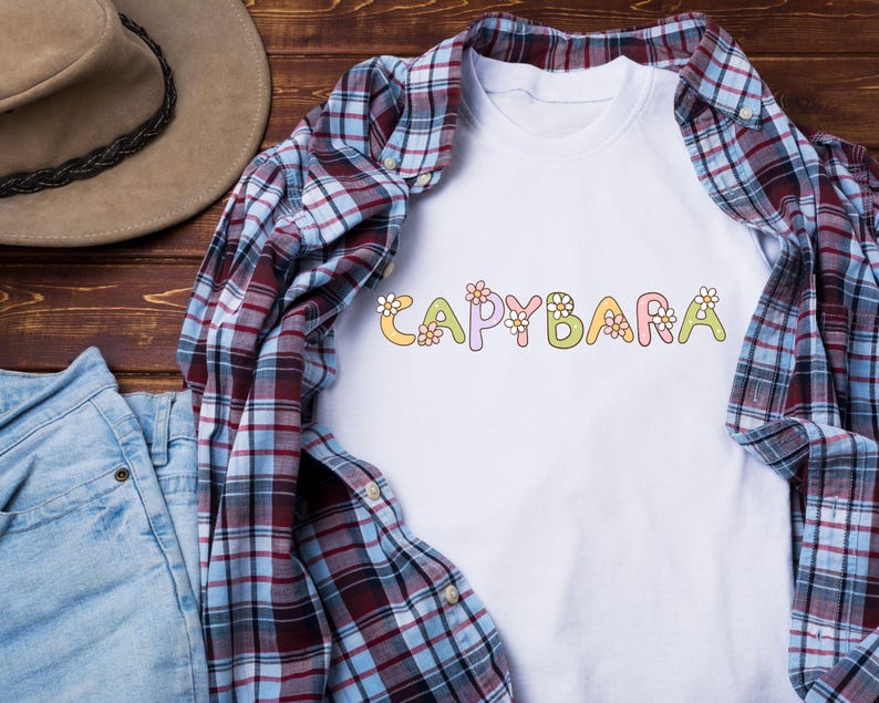 May include: White t-shirt with the word "CAPYBARA" in colorful, floral-decorated letters. The shirt is layered with a red, blue, and white plaid button-down shirt and blue jeans. A brown hat is visible in the background.