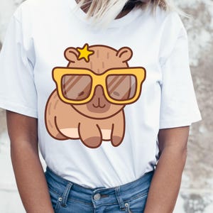 May include: White t-shirt featuring a cartoon capybara wearing yellow sunglasses and a star. The capybara is brown with a light brown belly. The sunglasses have brown lenses. The shirt is made of a soft material.