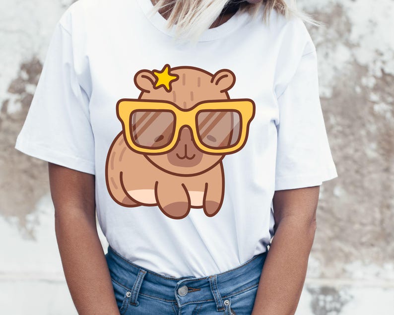 May include: White t-shirt featuring a cartoon capybara wearing yellow sunglasses and a star. The capybara is brown with a light brown belly. The sunglasses have brown lenses. The shirt is made of a soft material.