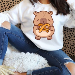 May include: White sweatshirt featuring a cartoon bear holding a pretzel. The bear is brown with a tan belly and the pretzel is golden brown. The sweatshirt is long-sleeved and the person is wearing blue jeans.