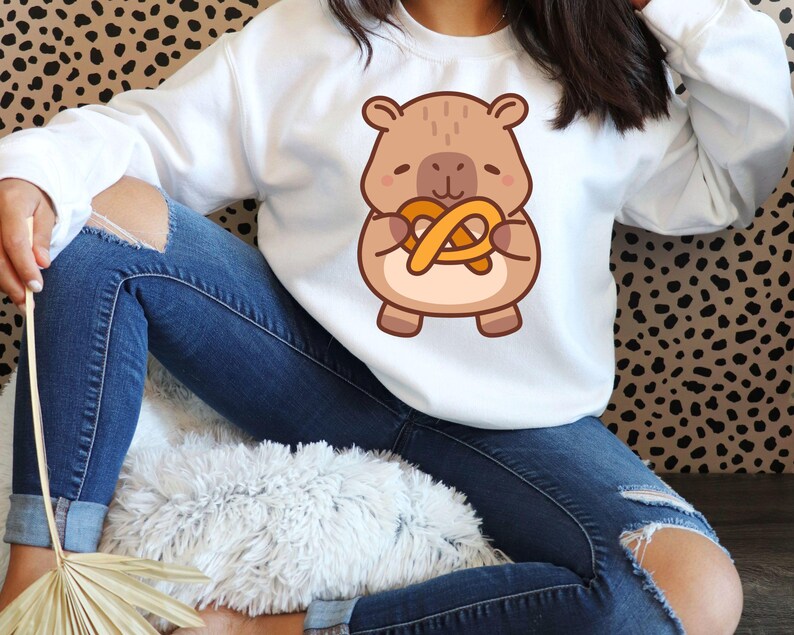 May include: White sweatshirt featuring a cartoon bear holding a pretzel. The bear is brown with a tan belly and the pretzel is golden brown. The sweatshirt is long-sleeved and the person is wearing blue jeans.
