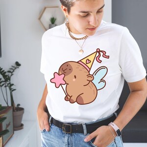 May include: White t-shirt featuring a cartoon illustration of a brown creature with a party hat, wings, and a pink star wand. The design is centered on the shirt. The person is wearing blue jeans and a black belt.