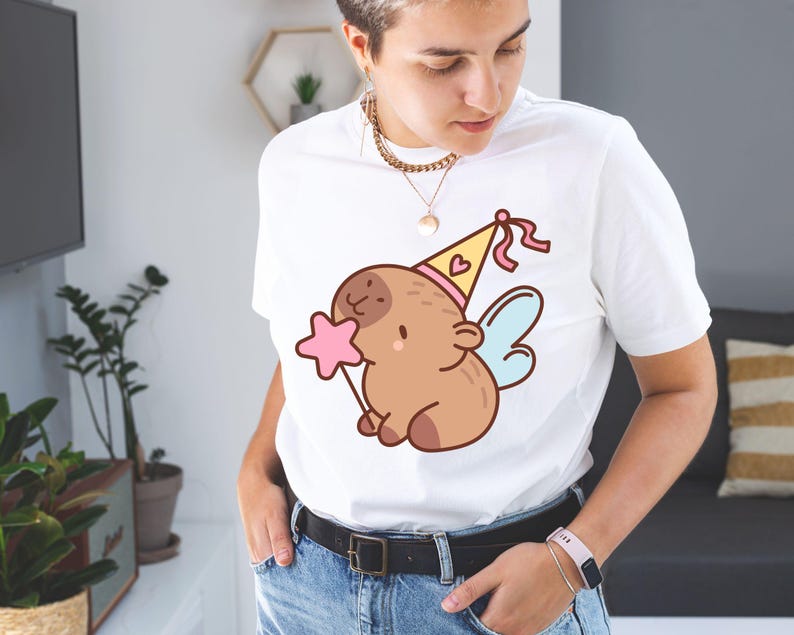 May include: White t-shirt featuring a cartoon illustration of a brown creature with a party hat, wings, and a pink star wand. The design is centered on the shirt. The person is wearing blue jeans and a black belt.