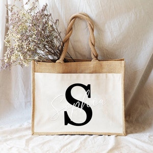 May include: A natural jute tote bag with a white canvas insert. The bag has rope handles and features a black letter "S" with the name "Sophia" written in cursive in white.