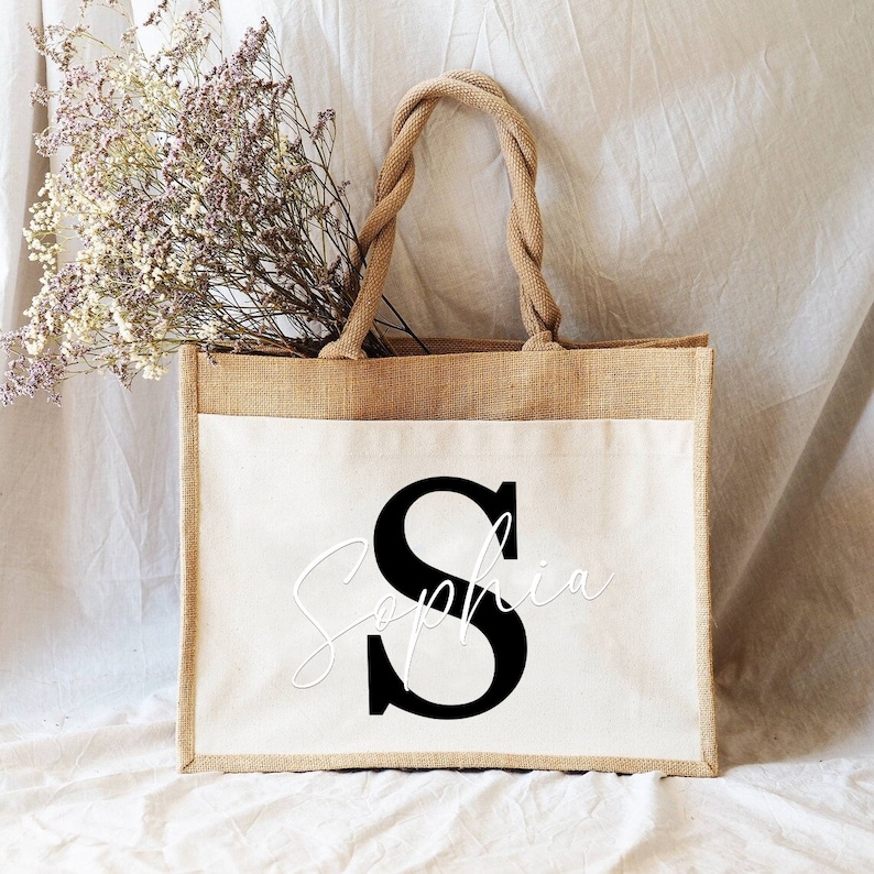 May include: A natural jute tote bag with a white canvas insert. The bag has rope handles and features a black letter "S" with the name "Sophia" written in cursive in white.