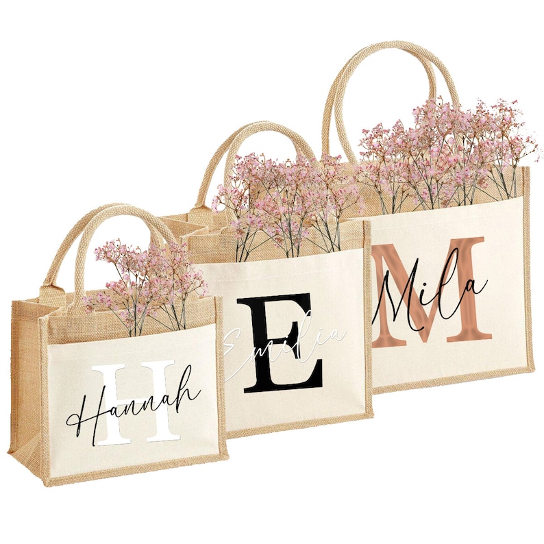 May include: Three burlap tote bags with personalized names and initial letters. The bags are filled with pink flowers and have a natural fabric texture.