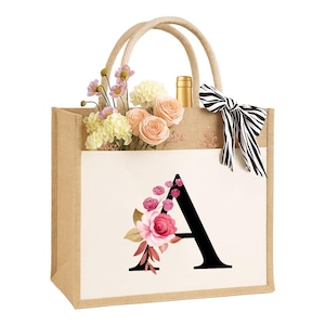 以下が含まれることがあります： ベージュのジュートトートバッグ。花の模様と「A」の文字がプリントされています。バッグにはロープの持ち手と、白と黒のストライプのリボンが付いています。バッグの中には花とワインボトルが入っています。