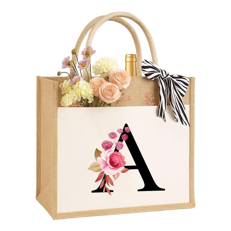 以下が含まれることがあります： ベージュのジュートトートバッグ。花の模様と「A」の文字がプリントされています。バッグにはロープの持ち手と、白と黒のストライプのリボンが付いています。バッグの中には花とワインボトルが入っています。