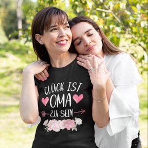 Könnte beinhalten: Ein schwarzes T-Shirt mit dem Text "Glück ist Oma zu sein" in weiß mit zwei rosa Herzen und einem rosa Pfeil. Das Shirt wird von einer Frau getragen, die eine andere Frau umarmt.