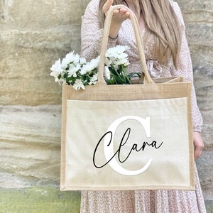 May include: A beige jute tote bag with a white canvas pocket featuring the name "Clara" in a black cursive font with a large white "C" initial.