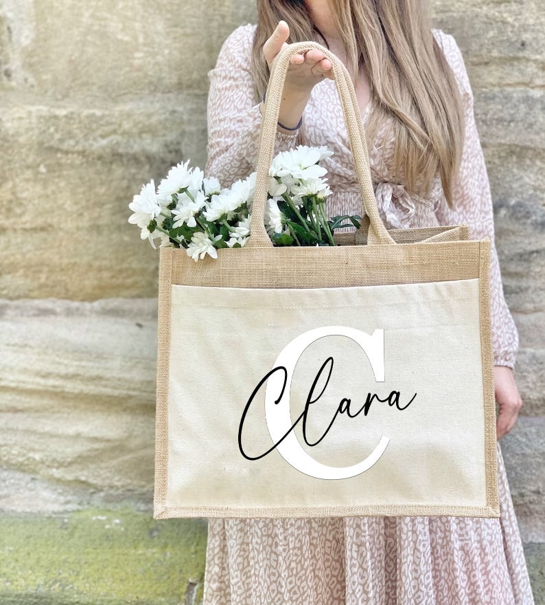 May include: A beige jute tote bag with a white canvas pocket featuring the name "Clara" in a black cursive font with a large white "C" initial.