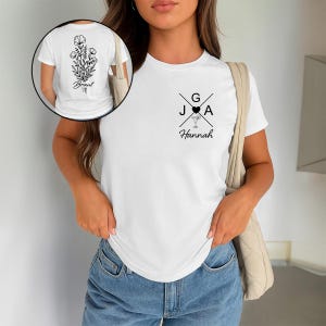 Op de afbeelding: Wit T-shirt met een zwart bloemmotief op de rug en een ontwerp op de voorkant met de letters G, J en A, een hart en een cocktailglas. De naam "Hannah" staat eronder gedrukt. Het T-shirt wordt door een persoon gedragen.