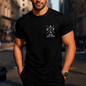 Könnte beinhalten: Schwarzes T-Shirt mit weißem Aufdruck auf der Brust. Der Aufdruck zeigt die Buchstaben "A" und "P" in einem Kreuz-Design, mit dem Namen "Noah" und dem Jahr "EST. 2026" darunter. Das T-Shirt hat kurze Ärmel und einen Rundhalsausschnitt.