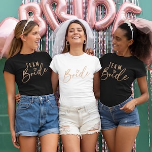 Op de afbeelding: Drie vriendinnen dragen bijpassende shirts met de tekst "Team Bride" in gouden letters. De shirts zijn zwart, wit en zwart. De vriendinnen lachen en poseren voor een groene muur met roze ballonnen die "Bride" spellen.