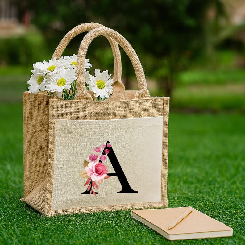 以下が含まれることがあります： ピンクの花で飾られた黒い文字「A」が特徴のクリーム色のポケットが付いたジュートトートバッグ。バッグには白いデイジーが入っており、タン色のハンドルが付いています。小さなノートと鉛筆が草の上にあります。
