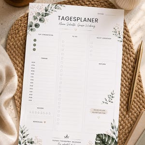 Könnte beinhalten: Ein weißer Tagesplaner mit grünen botanischen Akzenten und dem Text "TAGESPLANER". Der Planer enthält Abschnitte für Prioritäten, To-Do-Listen, Notizen und einen Zeitplan. Ein goldener Stift und eine Tasse Kaffee sind ebenfalls sichtbar.