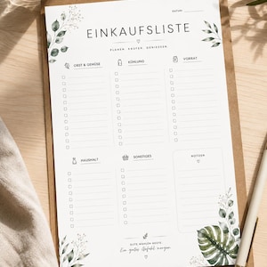 Könnte beinhalten: Einkaufszettel-Notizblock mit Kraftpapier-Rückseite. Das weiße Papier hat Abschnitte für Lebensmittel, Haushaltsartikel und Notizen mit dekorativen botanischen Illustrationen. Der Text "EINKAUFSLISTE" steht oben.