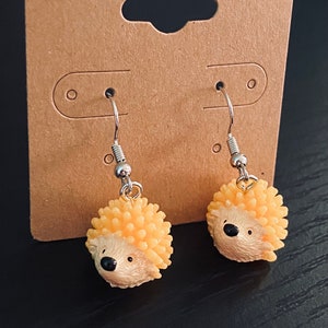 Peut inclure: Une paire de boucles d'oreilles pendantes argentées avec un charme mignon de hérisson jaune. Le hérisson a des yeux noirs et un petit nez noir.