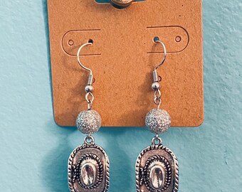 May include: Silver-toned cowboy hat earrings with a textured bead and a decorative rope border. The earrings hang from silver-colored fish hook ear wires. The earrings are displayed on a brown earring card.