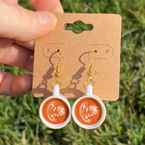 May include: A pair of coffee-themed earrings. Each earring features a white cup with a handle and a brown coffee design. The earrings hang from gold-colored hooks and are displayed on a brown card.