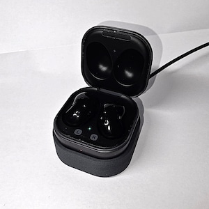 Könnte beinhalten: Schwarze kabellose Ohrhörer in einem Ladeetui. Das Etui ist geöffnet und zeigt die Ohrhörer und die Ladeschlitze. Das Etui ist mit einem schwarzen Kabel verbunden. Die Ohrhörer sind mit "L" und "R" gekennzeichnet.
