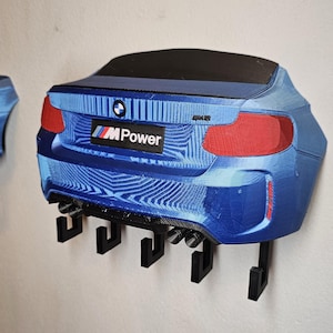 May include: Two blue car-shaped key holders fixed to a wall. The key holders are designed to resemble the rear of a car, with red tail lights and a black bumper. Each key holder has several black hooks for keys. The BMW M Power logo is visible.