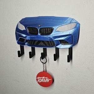 Puede incluir: Un llavero azul con forma de coche con cuatro ganchos negros. El llavero está montado en una pared blanca. Una etiqueta circular roja con la palabra "treaw" en escritura blanca cuelga de uno de los ganchos.