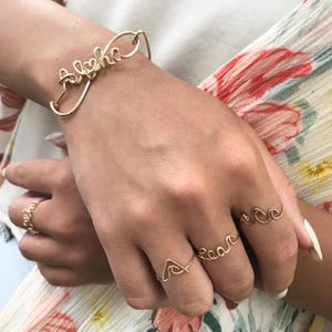 Puede incluir: Conjunto de joyas de alambre dorado con una pulsera con la palabra "Aloha" y tres anillos con las palabras "Peace", "Love" y un diseño de ola.