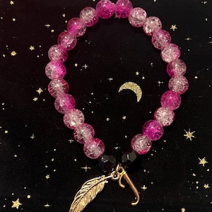 Puede incluir: Un brazalete de cuentas de cristal rosa y blanco con un dije de pluma dorada y un dije de letra J dorada. El brazalete está sobre un fondo negro con una luna creciente.