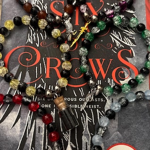 Peut inclure: Plusieurs bracelets de perles de différentes couleurs, dont le noir, le rouge, le jaune et le vert, avec des breloques argentées. Les bracelets sont présentés sur un livre intitulé "Six of Crows" de Leigh Bardugo, avec un autocollant Netflix.