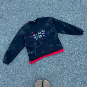 May include: Black crew neck sweatshirt with the word "BUF" embroidered in white, red, and blue. The sweatshirt has a red band at the bottom and a tie-dye effect. The image also shows a pair of brown clogs.