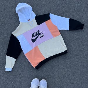 May include: A color-block Nike SB hoodie in cream, black, white, and peach. The front features a light purple panel with the Nike SB logo in black. The hoodie has a kangaroo pocket and a white hood.