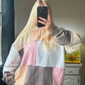 May include: A color-block hoodie in a patchwork design. The hoodie features panels of beige, pink, white, and brown. A small Nike logo is visible on the chest. The sleeves are a darker shade of brown and gray.